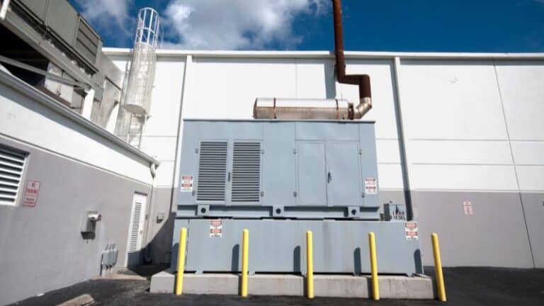 Outdoor installation of a large industrial generator with a gray enclosure. The generator is equipped with an exhaust pipe and is surrounded by yellow safety poles. The setting appears to be a facility with a white building in the background.