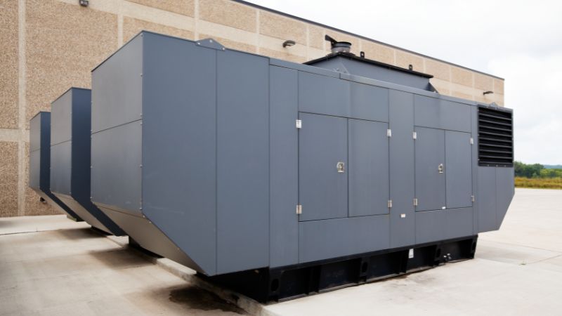 three industrial emergency power standby generators Three sizable, stationary standby generators painted in gray, positioned outdoors next to a building.