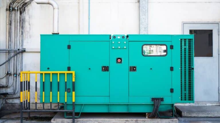 Large green industrial generator on a concrete platform outside, surrounded by pipes and cables.