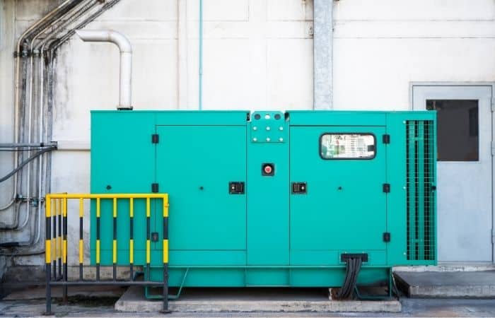 Large green industrial generator on a concrete pad outside a building, with a door and small yellow safety barrier in front.