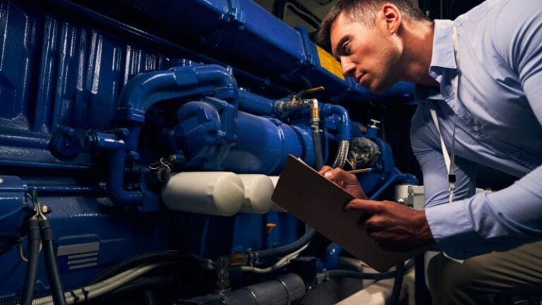 Man in blue shirt checks industrial machinery, taking notes on a clipboard while examining blue mechanical parts.