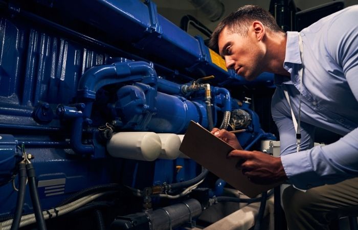A man in a dress shirt examines machinery and takes notes on a clipboard.
