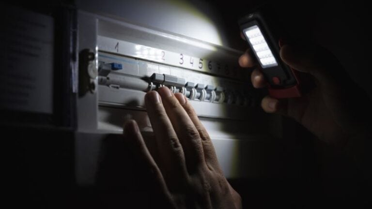 A person inspects an electrical breaker panel with a flashlight in a dimly lit room.