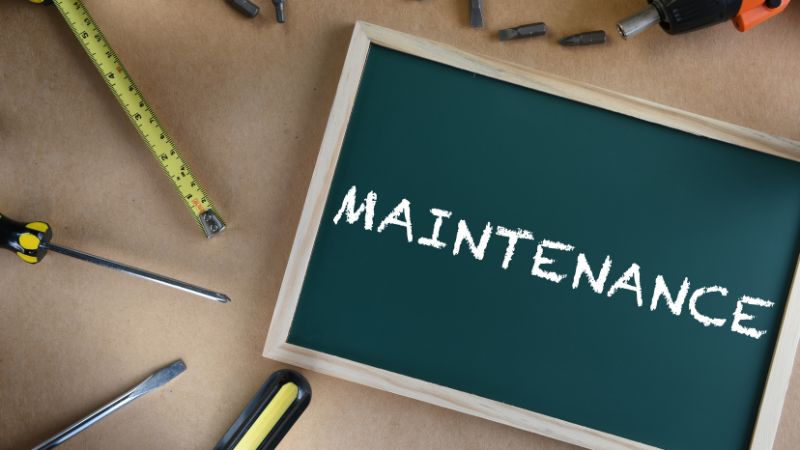 Chalkboard with the word "MAINTENANCE" written on it, surrounded by tools like screwdrivers, a measuring tape, and a utility knife on a brown surface.