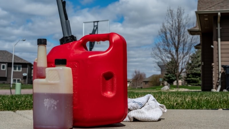 Gasoline containers on driveway with house in background, outdoor maintenance equipment, safety storage, home repair tools, lawn care supplies.